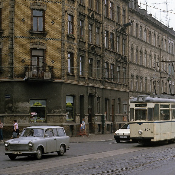 A street lined with brick buildings, with a tram and a couple old cars in front of it.